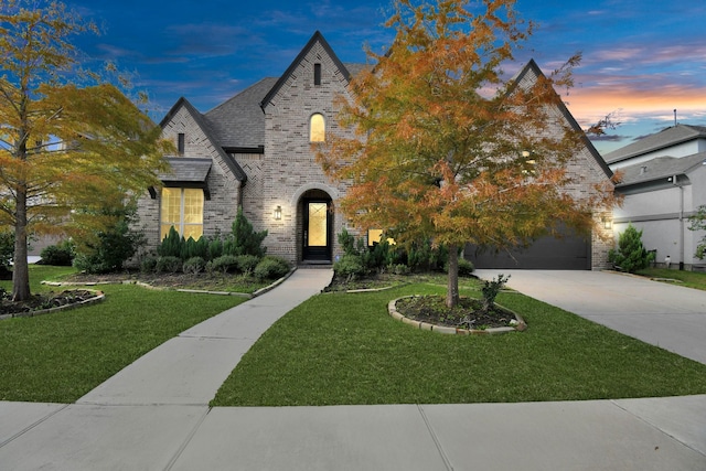 french country inspired facade with brick siding, a yard, a shingled roof, a garage, and driveway