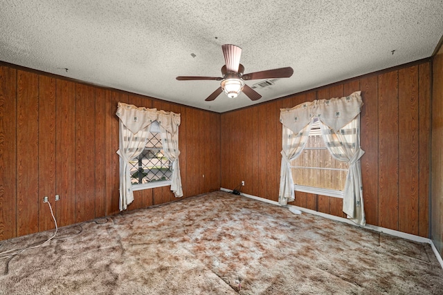 carpeted spare room with a textured ceiling, visible vents, a ceiling fan, and wooden walls