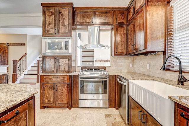 kitchen with stainless steel appliances, tasteful backsplash, a barn door, a sink, and wall chimney range hood