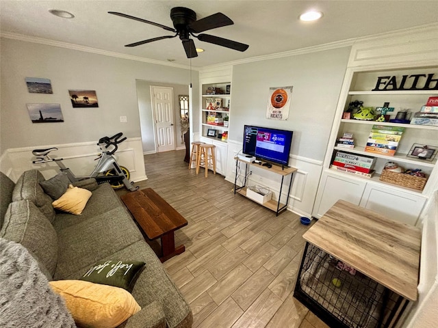 living area featuring built in features, a ceiling fan, a wainscoted wall, crown molding, and light wood-type flooring