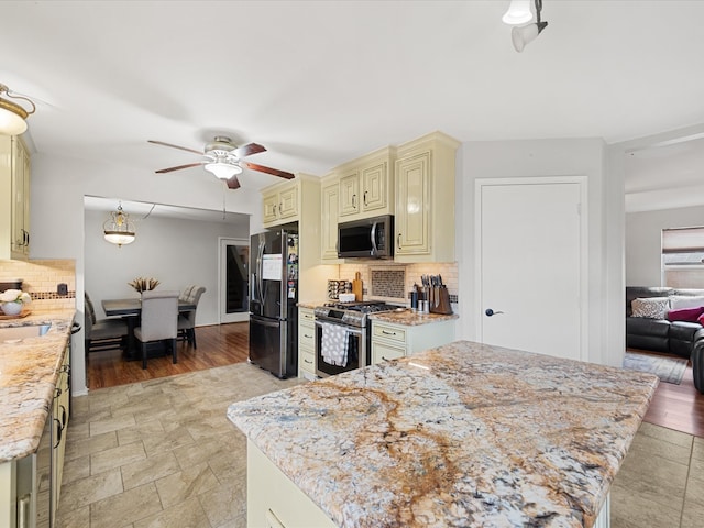 kitchen featuring cream cabinetry, a ceiling fan, light stone counters, appliances with stainless steel finishes, and decorative backsplash