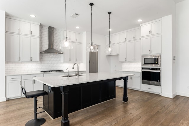 kitchen with a center island with sink, wall chimney exhaust hood, appliances with stainless steel finishes, a sink, and a kitchen breakfast bar