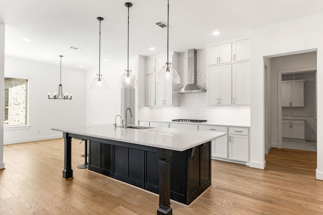 kitchen featuring light stone counters, decorative light fixtures, a kitchen island with sink, a sink, and wall chimney exhaust hood