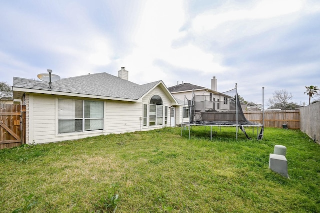 back of house with a yard, a trampoline, a fenced backyard, and roof with shingles