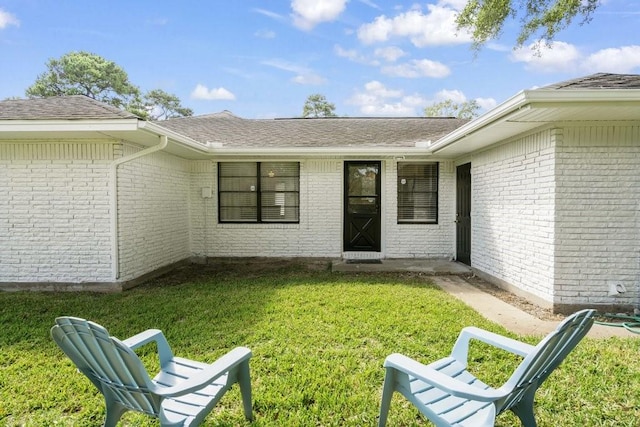 doorway to property with an attached garage, roof with shingles, a yard, and brick siding