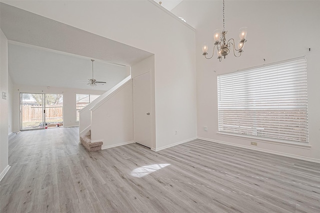 unfurnished living room with high vaulted ceiling, light wood-style flooring, ceiling fan with notable chandelier, baseboards, and stairs