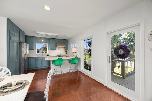 kitchen with dark wood finished floors, light countertops, wall chimney range hood, a peninsula, and stainless steel fridge with ice dispenser