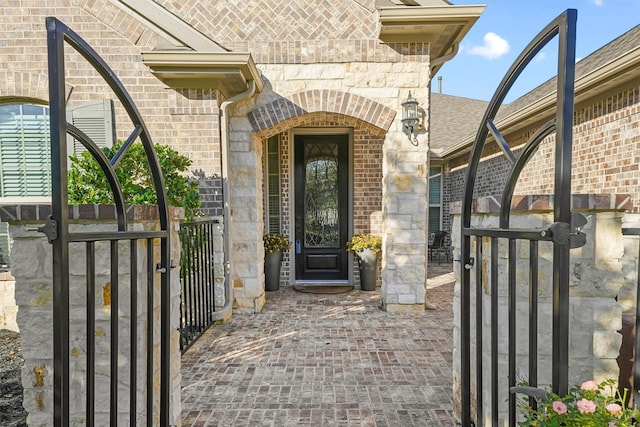 view of exterior entry with brick siding, a shingled roof, and a gate