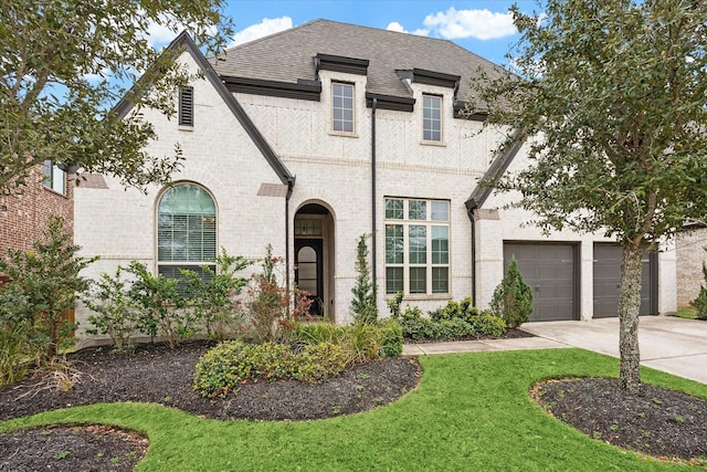 french provincial home featuring driveway, a front lawn, roof with shingles, an attached garage, and brick siding