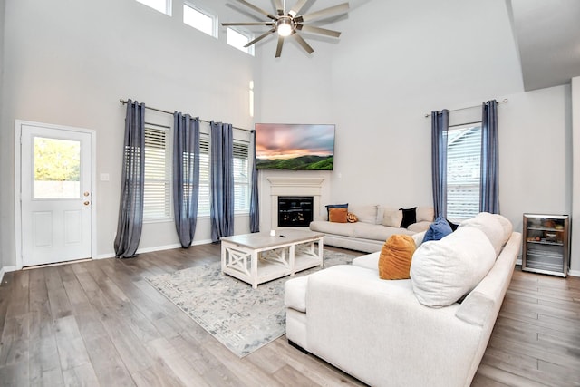 living area featuring a high ceiling, a ceiling fan, a glass covered fireplace, wood finished floors, and baseboards