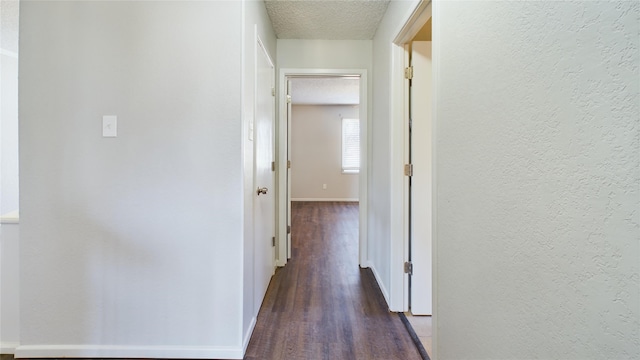 hallway featuring a textured ceiling, a textured wall, dark wood finished floors, and baseboards