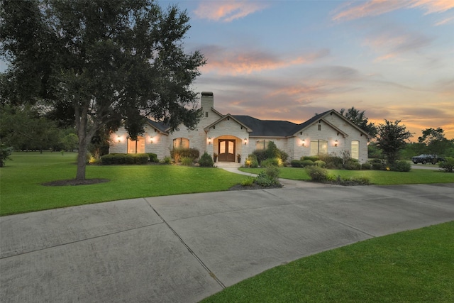 french provincial home featuring stone siding, a chimney, a lawn, and concrete driveway