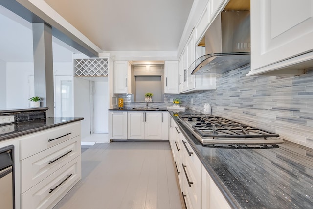 kitchen featuring a sink, white cabinets, wall chimney range hood, backsplash, and dark stone counters