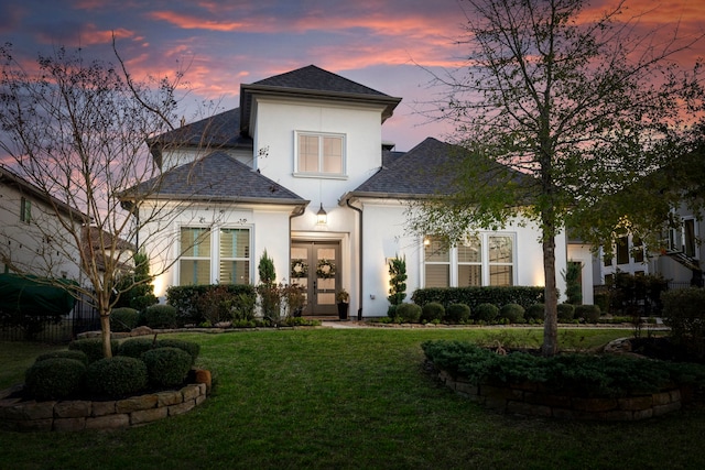 view of front of house featuring stucco siding, roof with shingles, a lawn, and french doors