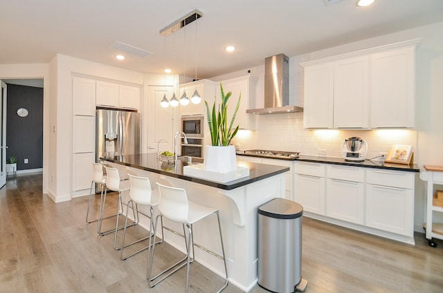 kitchen featuring stainless steel appliances, white cabinets, wall chimney range hood, an island with sink, and decorative light fixtures