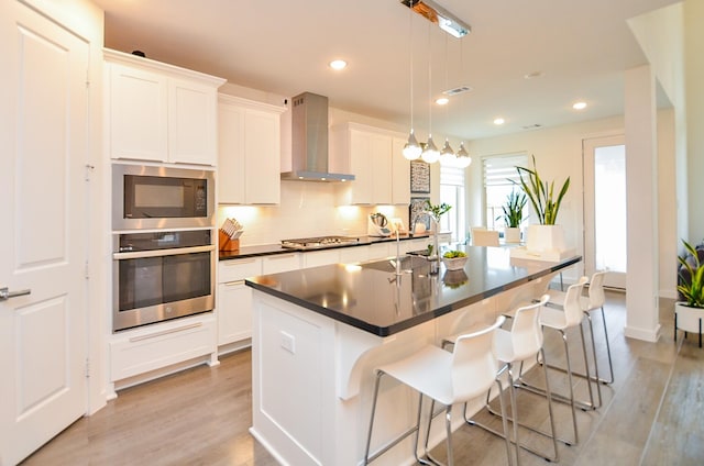 kitchen with dark countertops, wall chimney exhaust hood, a kitchen island with sink, stainless steel appliances, and pendant lighting