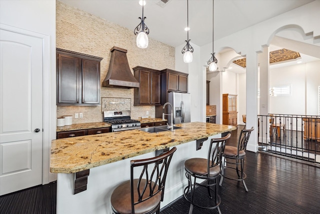 kitchen with stainless steel appliances, a sink, dark brown cabinets, custom exhaust hood, and a kitchen bar