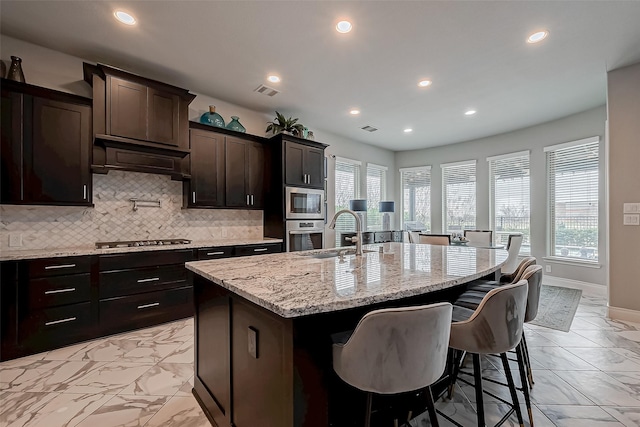 kitchen featuring a kitchen island with sink, stainless steel appliances, a sink, decorative backsplash, and light stone countertops