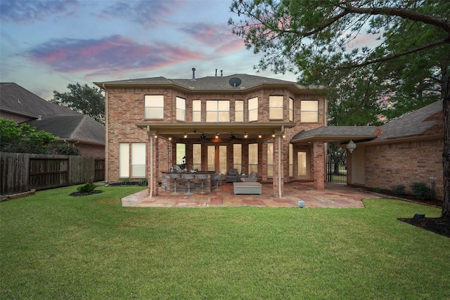 back of property at dusk featuring a lawn, ceiling fan, fence, a patio area, and brick siding