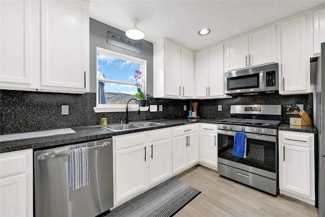 kitchen with stainless steel appliances, a sink, white cabinets, light wood finished floors, and dark stone countertops