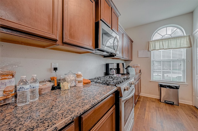 kitchen featuring baseboards, brown cabinetry, dark stone counters, light wood-style flooring, and appliances with stainless steel finishes