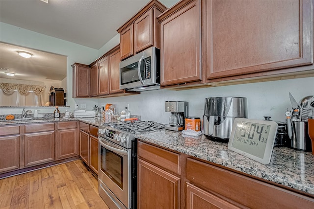 kitchen with stainless steel appliances, a sink, brown cabinets, light stone countertops, and light wood finished floors