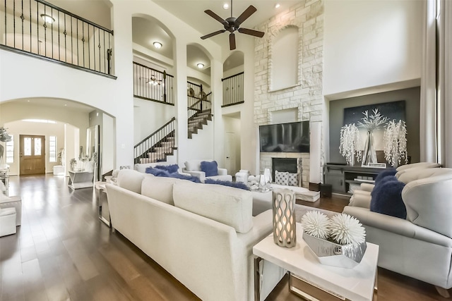 living room featuring arched walkways, dark wood finished floors, a towering ceiling, a stone fireplace, and stairs