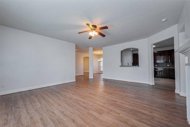 unfurnished living room with arched walkways, ceiling fan with notable chandelier, light wood-type flooring, and baseboards