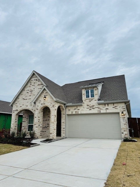 french country home with a garage, brick siding, concrete driveway, and roof with shingles