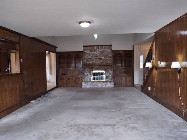 unfurnished living room featuring wooden walls, stairway, concrete flooring, and a brick fireplace