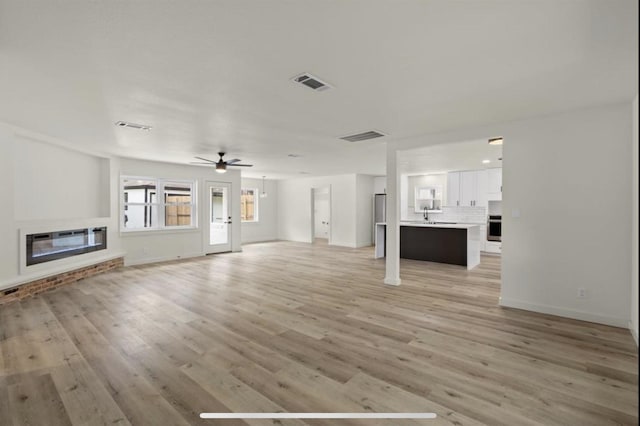 unfurnished living room featuring a sink, visible vents, a ceiling fan, light wood finished floors, and a glass covered fireplace