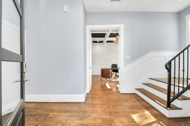 foyer entrance featuring a decorative wall, stairway, light wood-style floors, coffered ceiling, and beamed ceiling