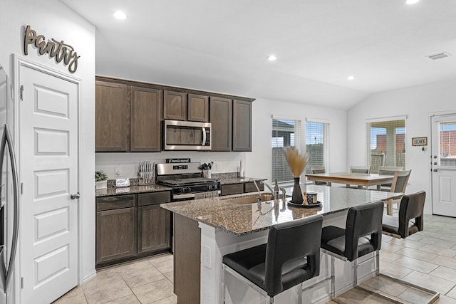 kitchen with dark brown cabinetry, an island with sink, light stone countertops, stainless steel appliances, and a sink