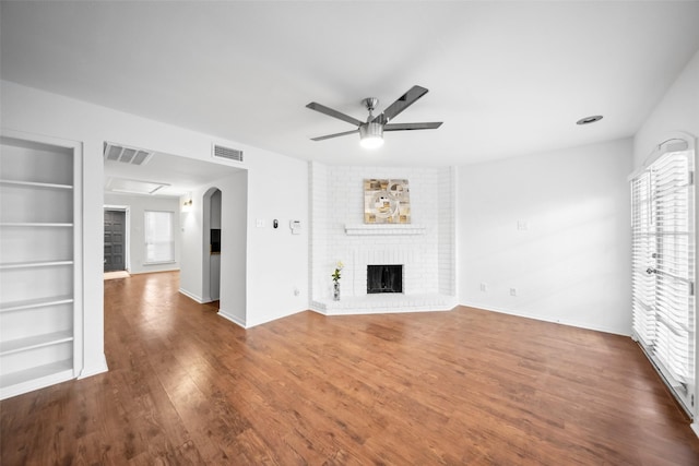 unfurnished living room featuring built in shelves, dark wood-style floors, visible vents, a brick fireplace, and ceiling fan