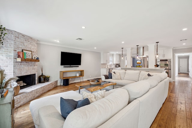 living room featuring light wood finished floors, visible vents, crown molding, a brick fireplace, and recessed lighting