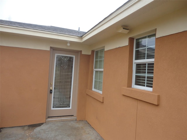 view of exterior entry featuring a shingled roof, a patio area, and stucco siding