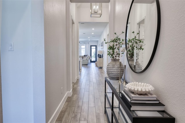 corridor featuring wood finished floors, a textured wall, baseboards, and a notable chandelier