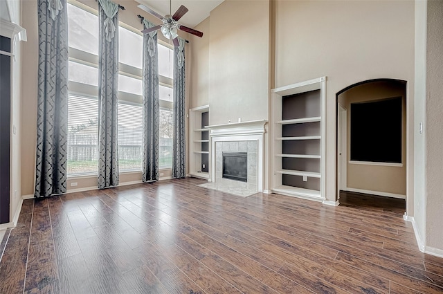 unfurnished living room with a high ceiling, dark wood-type flooring, ceiling fan, a tile fireplace, and baseboards