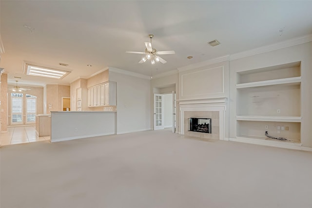 unfurnished living room with built in features, light colored carpet, a tiled fireplace, ornamental molding, and a ceiling fan