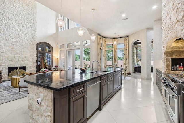 kitchen featuring arched walkways, stainless steel appliances, hanging light fixtures, a stone fireplace, and an island with sink