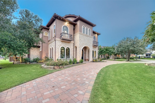 mediterranean / spanish-style home featuring a balcony, stone siding, a tiled roof, stucco siding, and a front lawn