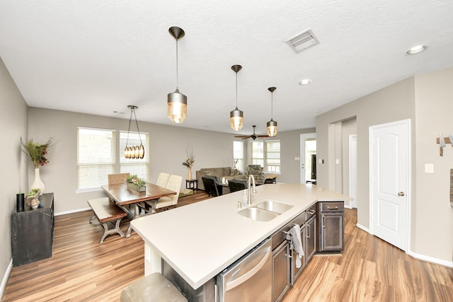 kitchen with hanging light fixtures, a sink, a kitchen island with sink, light countertops, and stainless steel dishwasher