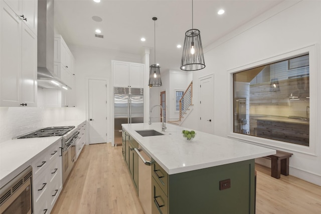 kitchen with built in appliances, a kitchen island with sink, white cabinets, wall chimney exhaust hood, and decorative light fixtures