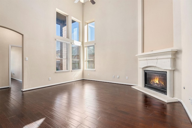 unfurnished living room with arched walkways, ceiling fan, dark wood finished floors, a towering ceiling, and a glass covered fireplace