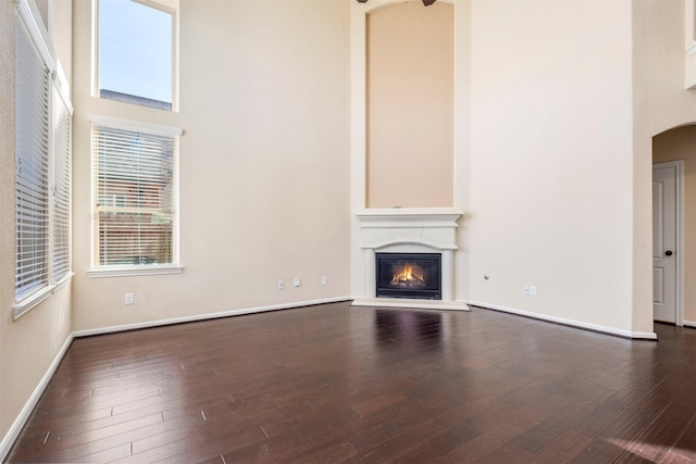 unfurnished living room featuring arched walkways, dark wood finished floors, a high ceiling, a glass covered fireplace, and baseboards