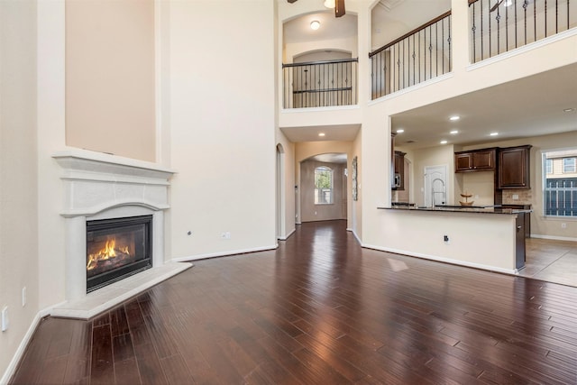 unfurnished living room featuring arched walkways, a towering ceiling, baseboards, dark wood-style floors, and a glass covered fireplace