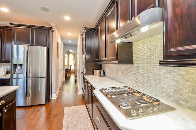 kitchen with arched walkways, dark wood finished floors, decorative backsplash, appliances with stainless steel finishes, and under cabinet range hood