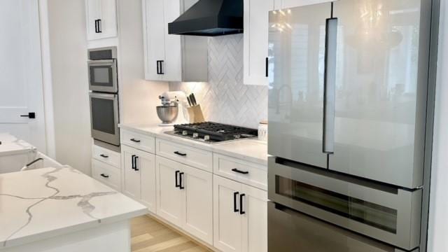 kitchen with appliances with stainless steel finishes, light stone counters, ventilation hood, white cabinetry, and backsplash