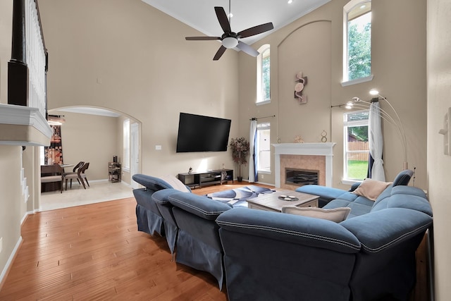 living area with ceiling fan, a tiled fireplace, arched walkways, light wood finished floors, and a high ceiling