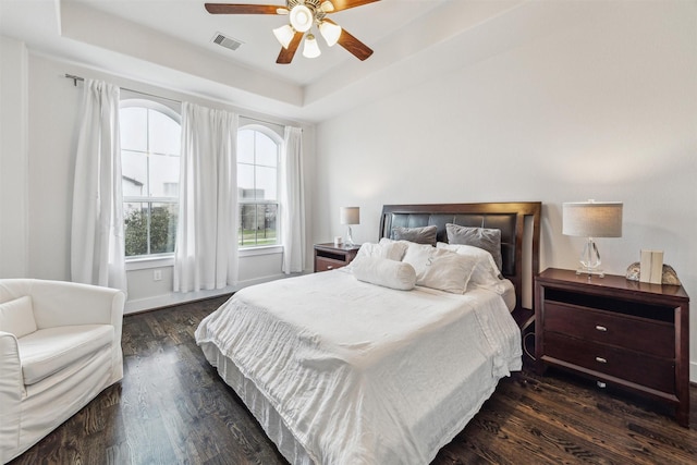 bedroom with a ceiling fan, a tray ceiling, visible vents, and dark wood finished floors